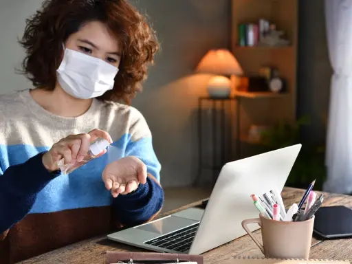 Kobieta w masce dezynfekuje dłonie płynem ze sprayu przy laptopie.