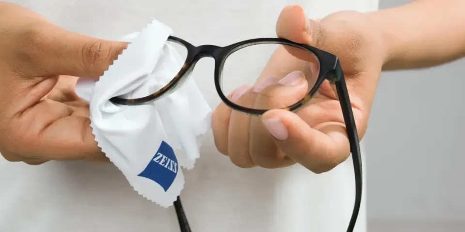 Dłonie czyszczą okulary białą ściereczką z logo Zeiss. Dowiedz się, czym czyścić okulary, by były idealnie przejrzyste.